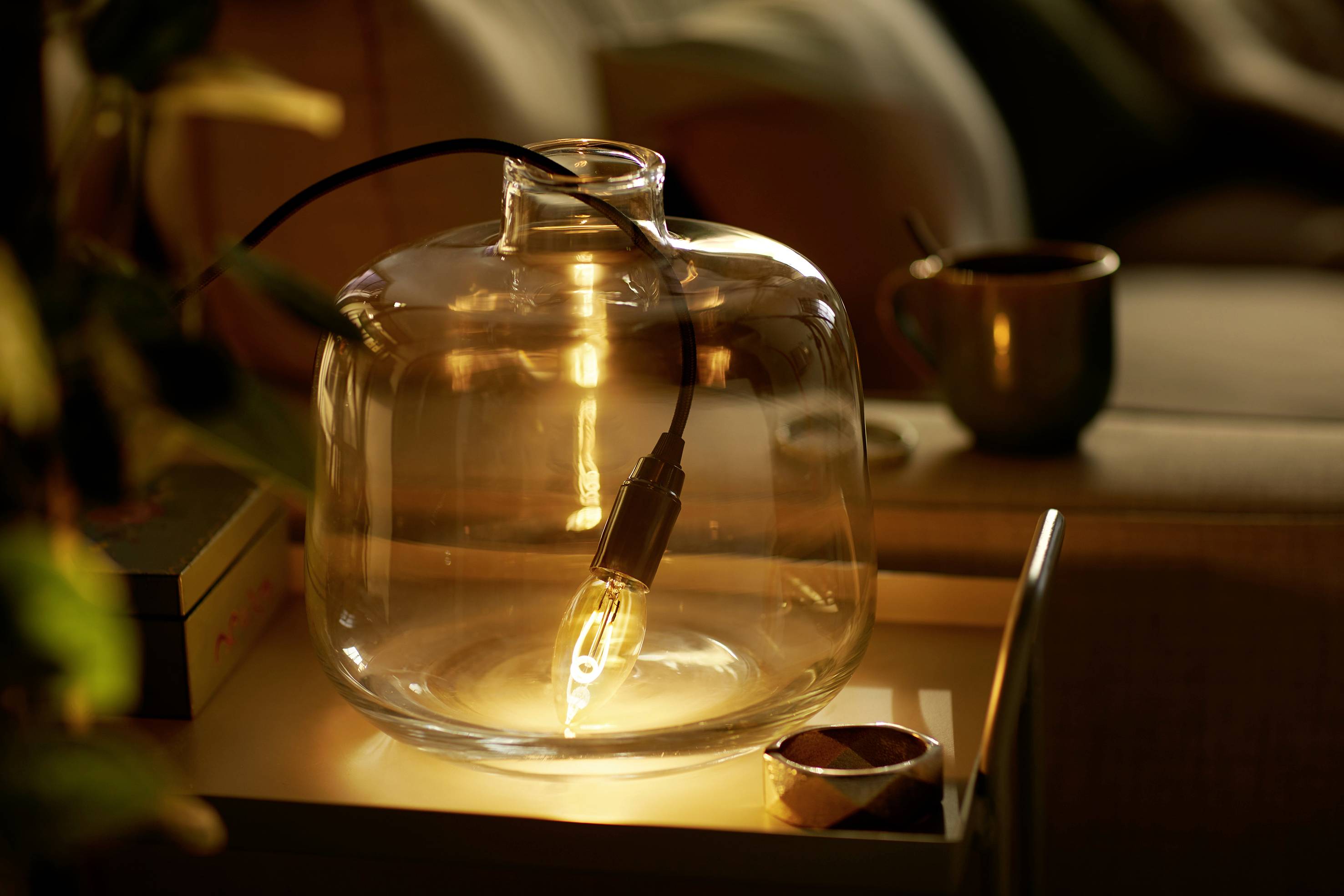 A light bulb in a large, clear glass container illuminates a table. A cup of coffee sits in the background.