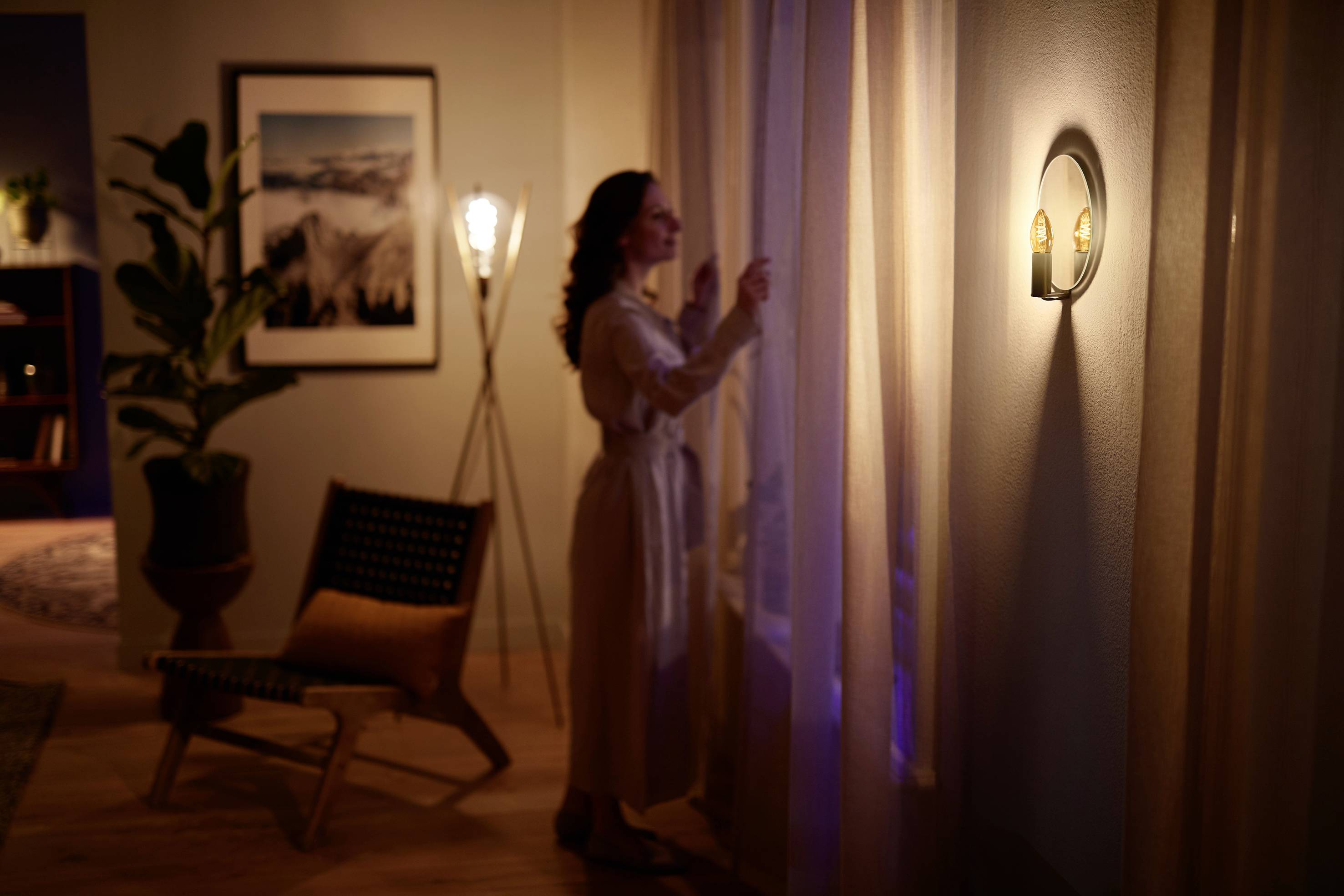 A woman in a dressing gown stands by the window in a cosily lit living room, drawing back the curtains.