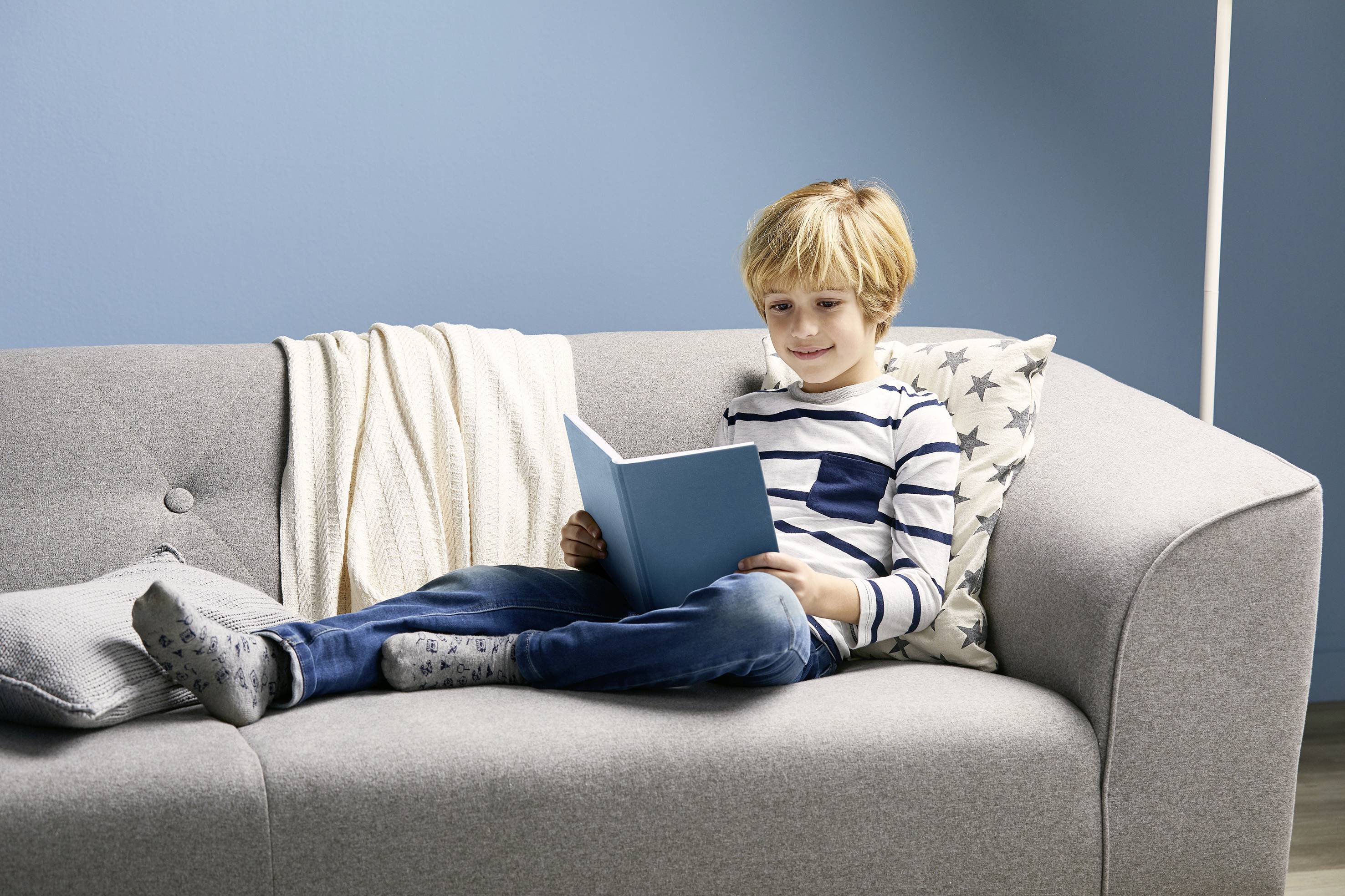 A boy is comfortably reading a blue book on a grey sofa. A light blue wall is visible in the background.