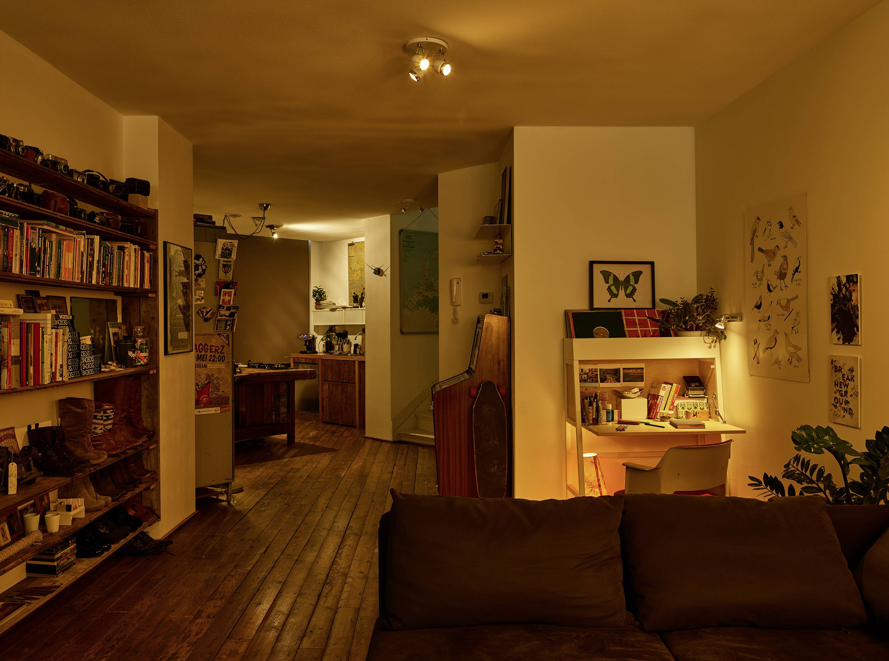 Cosy living room with warm lighting, shelves filled with books, a sofa and a desk with a lamp. Vintage decor and plants.