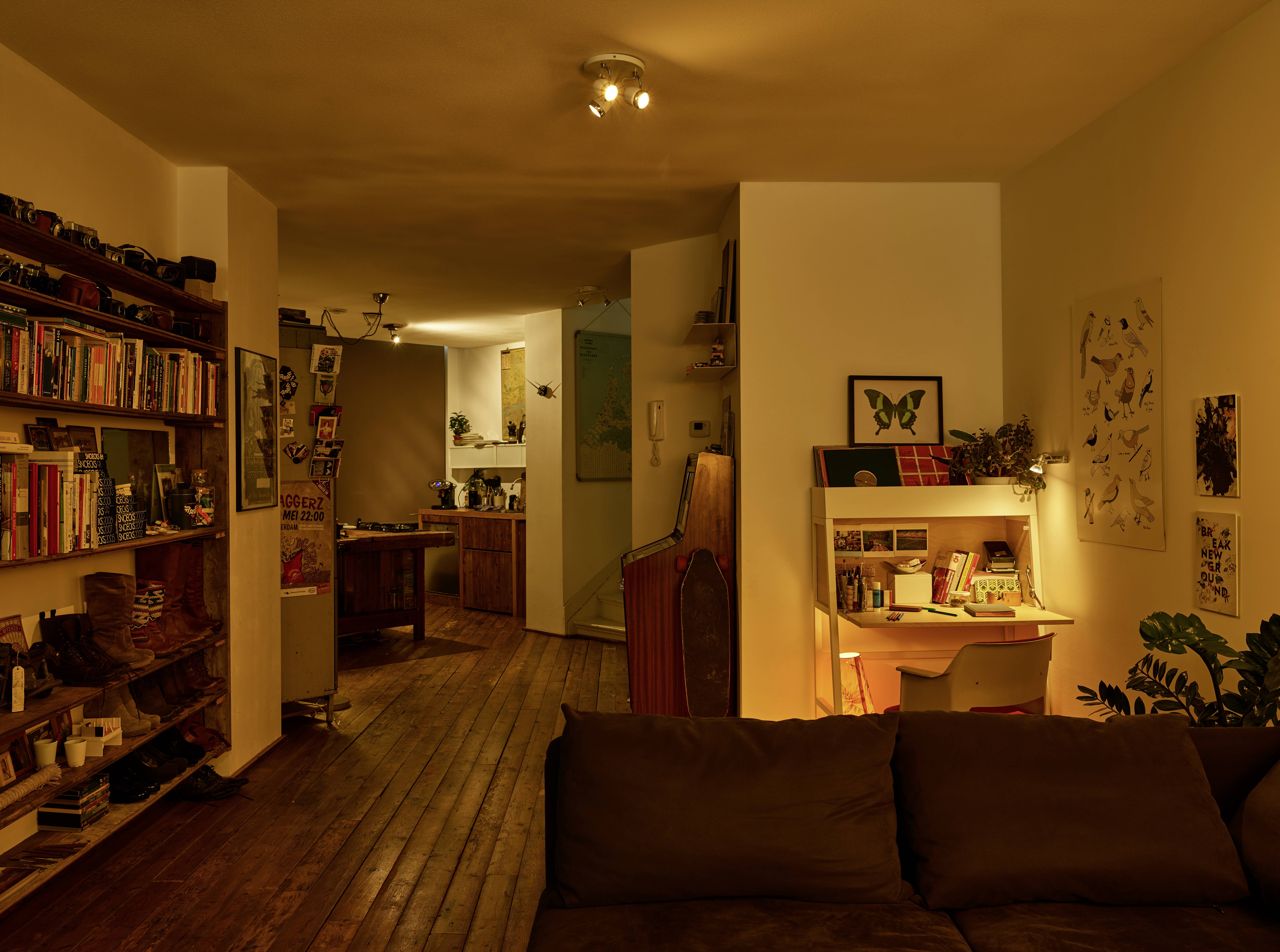 A cosy living room with wooden flooring and bookshelves. A kitchen is visible in the background. A desk corner with a lamp can be seen on the right.
