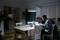 A man is working late at night on a laptop in a dimly lit room. A child is sitting on the sofa in the background.