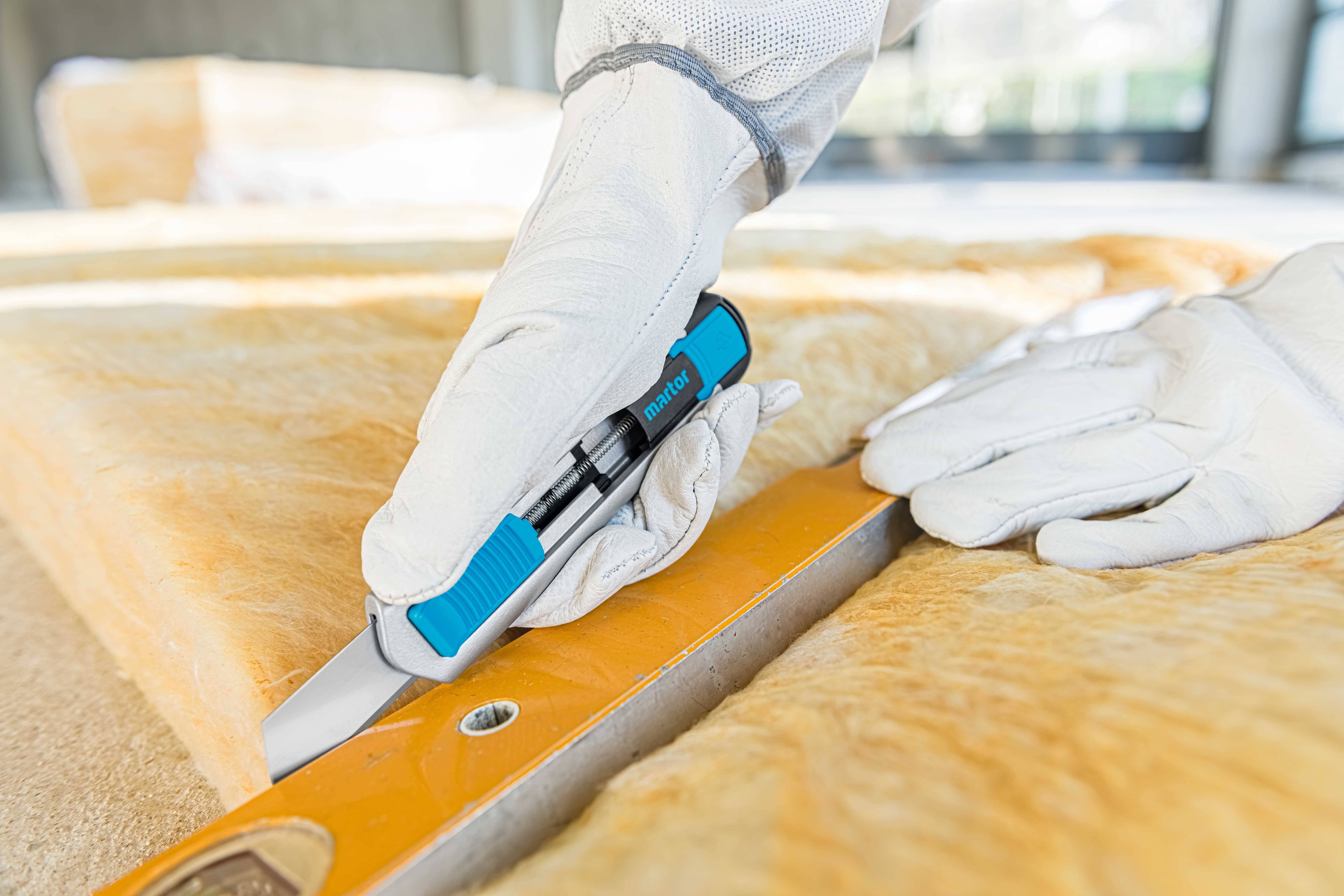 A person wearing gloves is precisely cutting a yellow insulation mat with a blue utility knife on a metal guide rail.