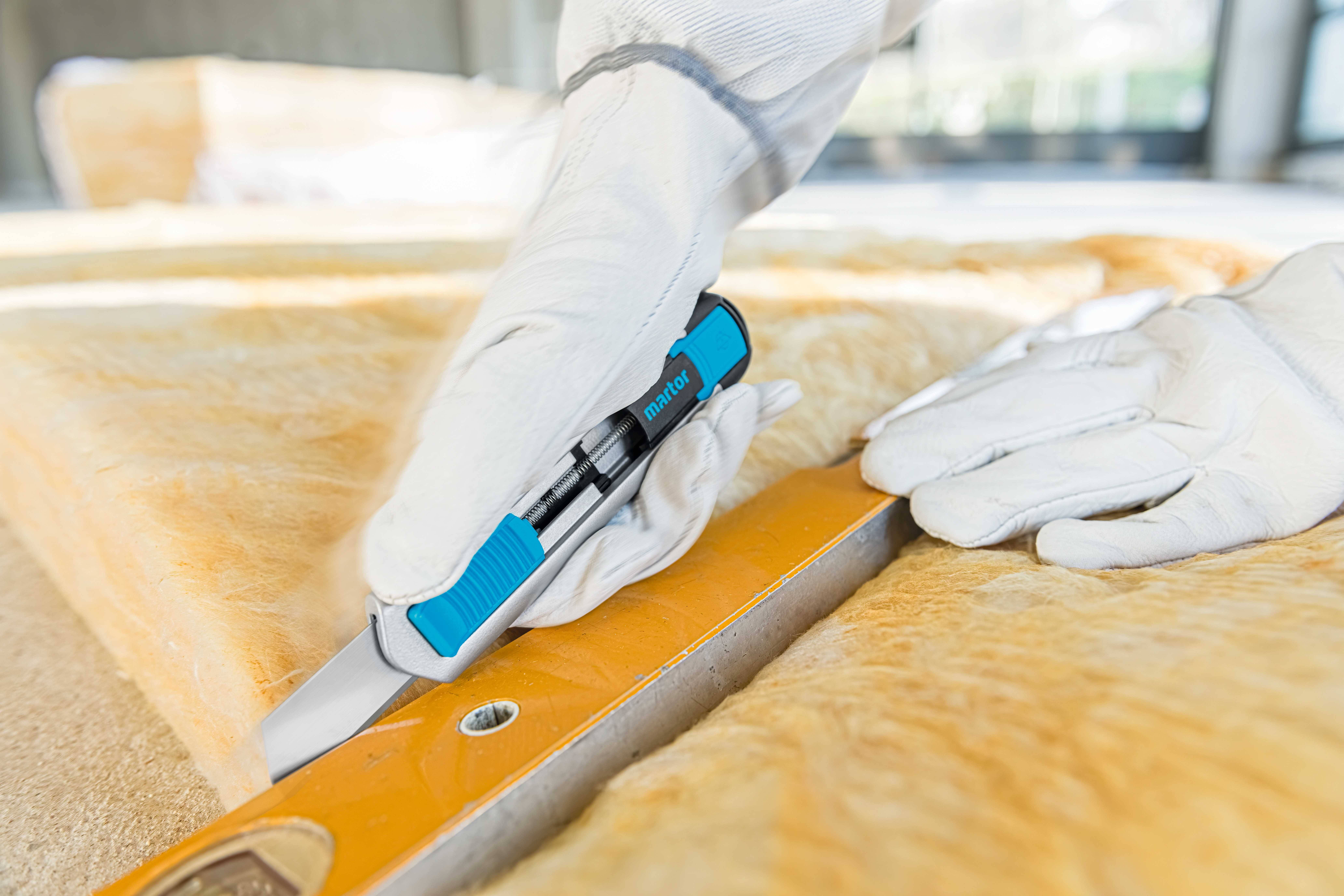 A person wearing white gloves is cutting glass wool with a blue knife along a ruler.