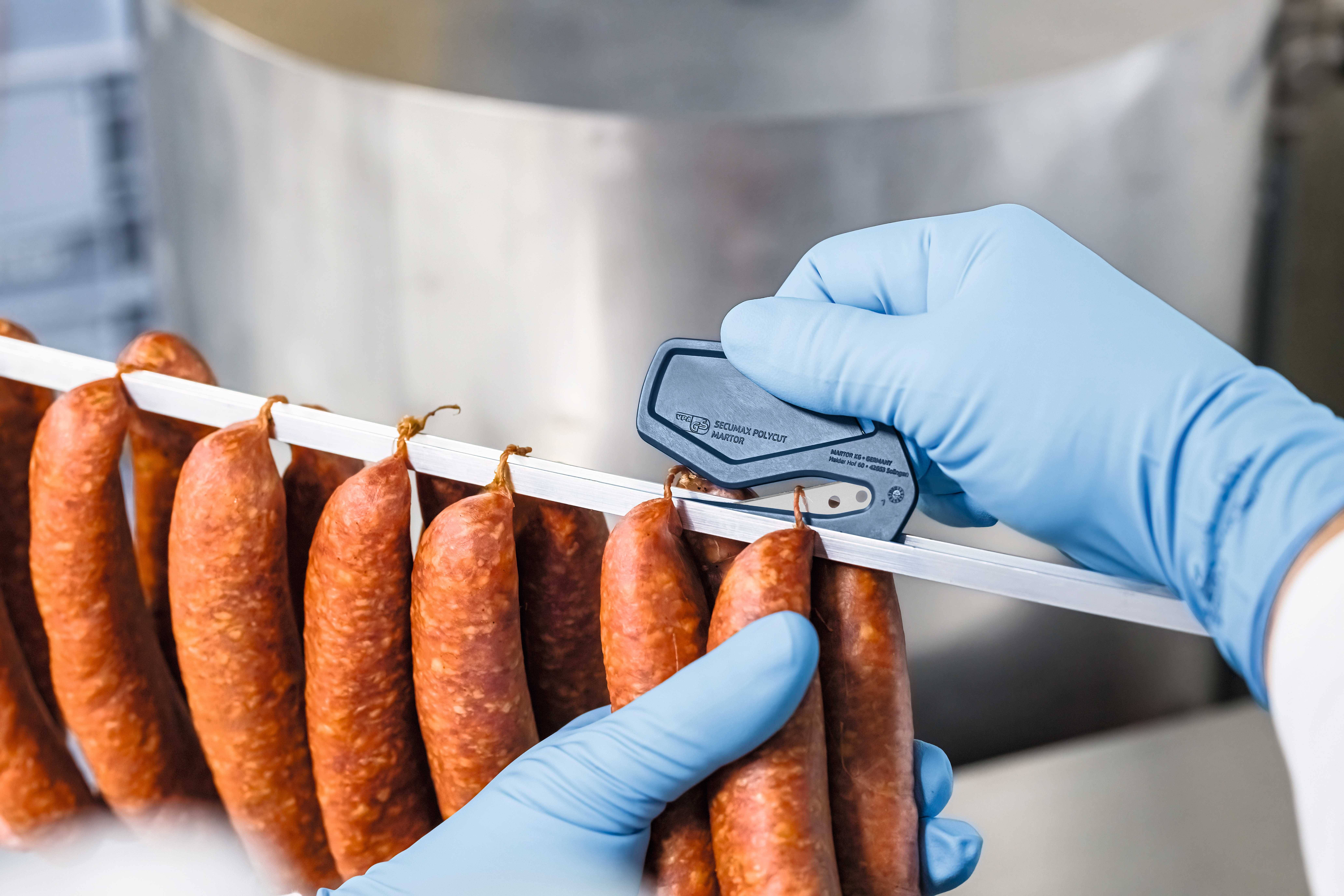 A person wearing blue gloves is measuring the diameter of hanging sausages in a production environment using a measuring instrument.