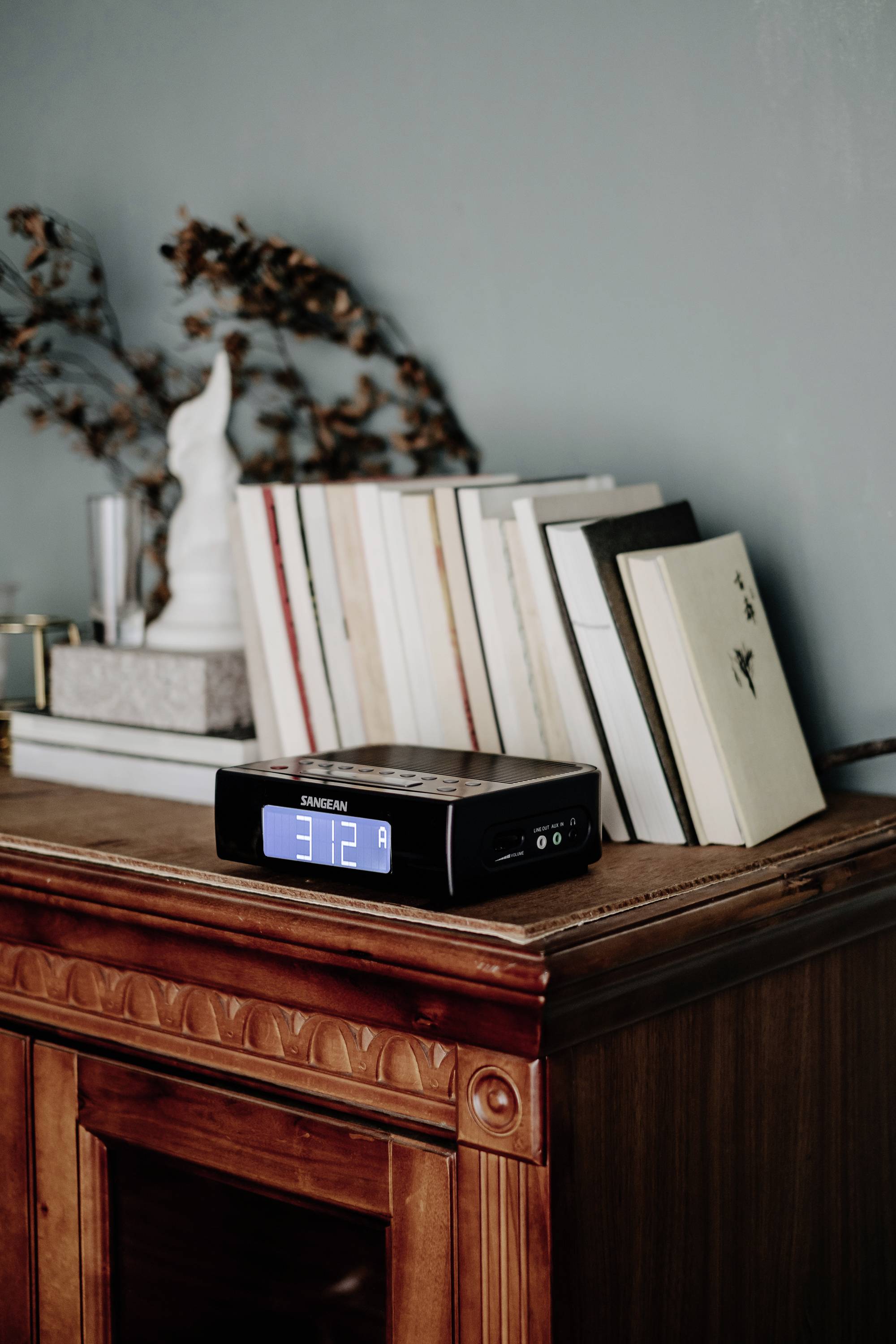 Digital alarm clock on a wooden table shows 3:12. Books are stacked at an angle in the background. A white candlestick serves as a decorative element.