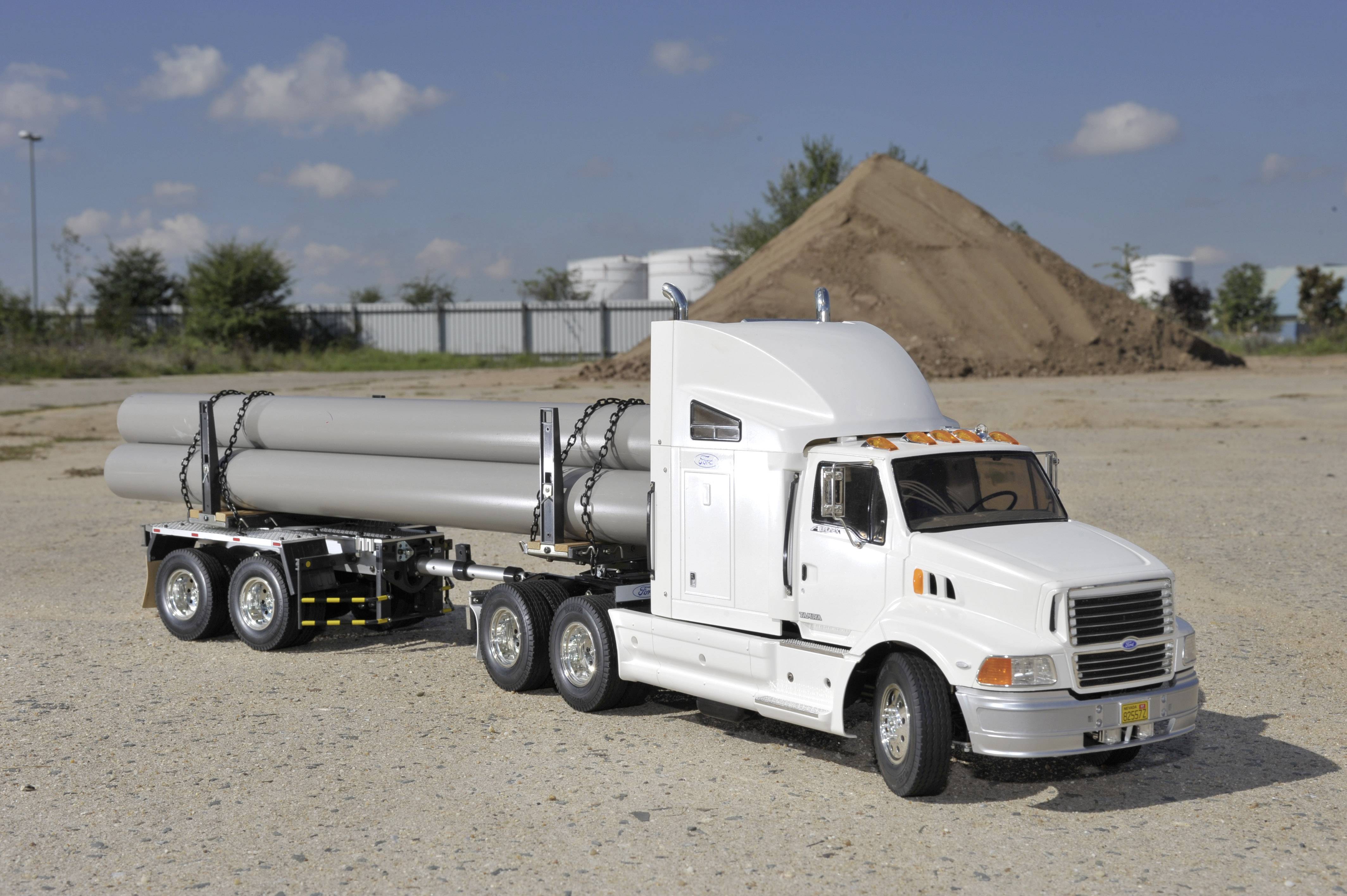 A white lorry is transporting long, grey pipes on a site with a gravel surface and an earth mound in the background.