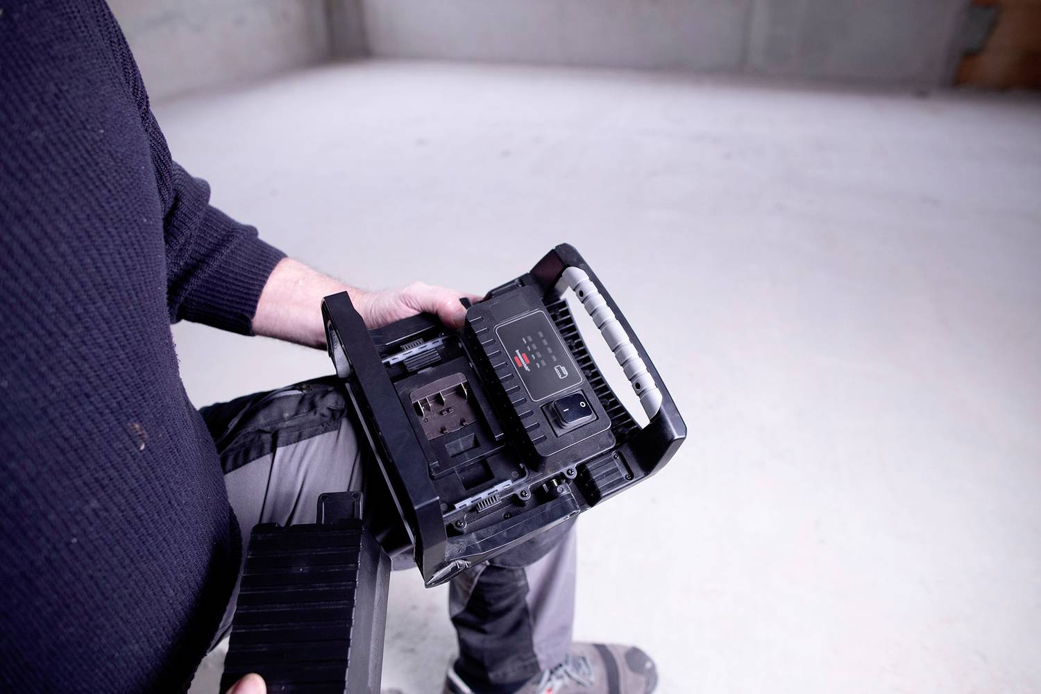 A person is holding a bagless handheld vacuum cleaner. The background is an empty room with a concrete floor.