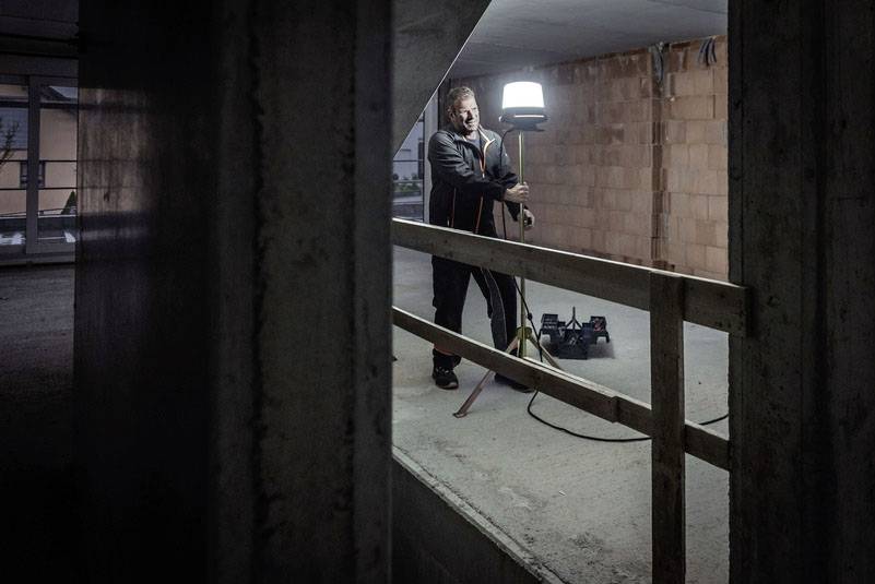 A man in an unfinished room stands next to a floor lamp on a tripod. Building materials and bare brick walls are in the background.