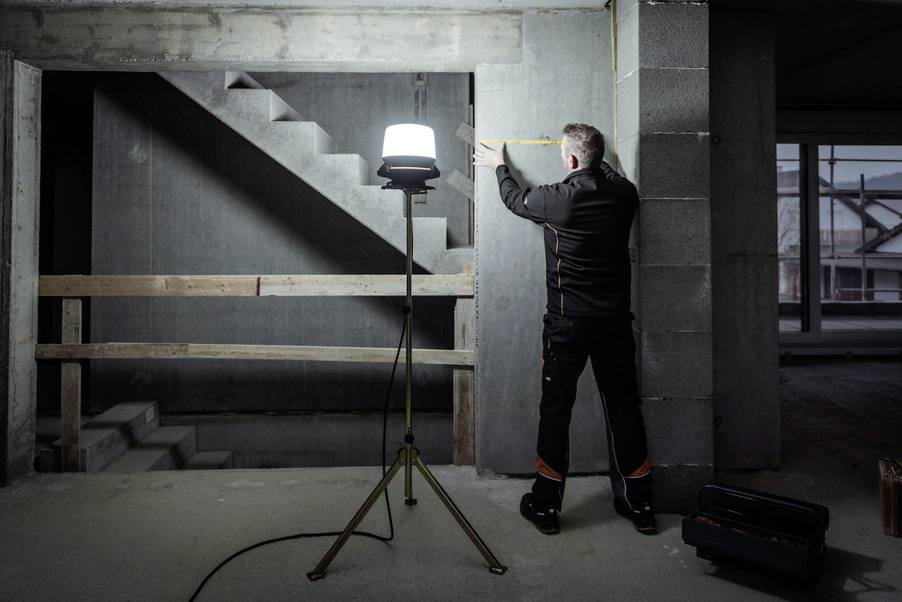 A person is measuring the wall in an unfinished building. A bright light stands on a tripod, illuminating the scene.