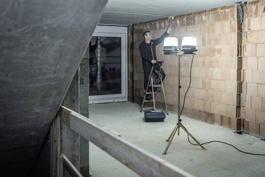 A worker stands on a ladder in an unfinished room, painting the ceiling. Two lamps illuminate the building site, with walls made of bricks.