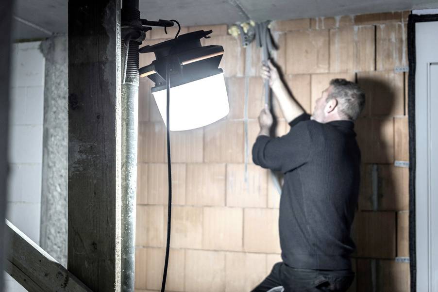 A man is working on a cable against an unplastered brick wall, illuminated by a bright construction site lamp.