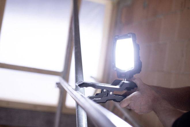 A person is holding a bright lamp, illuminating a piece of metal in an unfinished room. Background blurred, window visible.