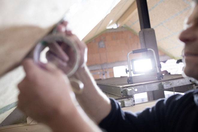A person is securing roof insulation with adhesive tape. A construction lamp and unfinished walls are visible in the background.