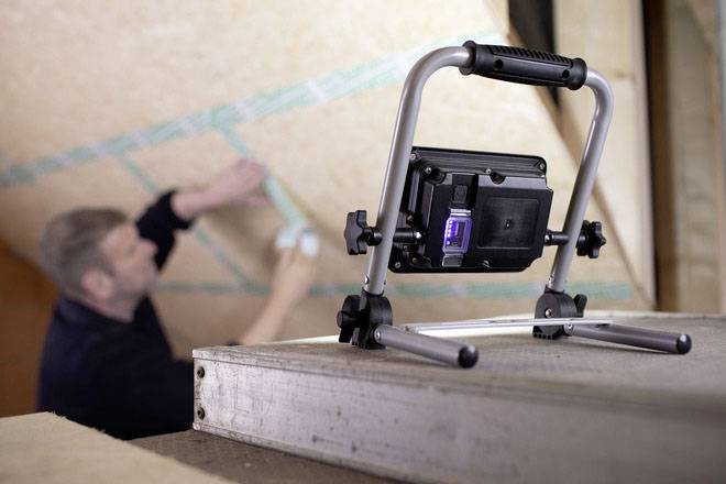 A person is working on a wall, with an LED work light on a stand in the foreground.