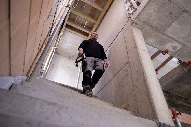 A worker in a black jumper and grey trousers is walking down a construction site staircase, holding a tool in his hand.
