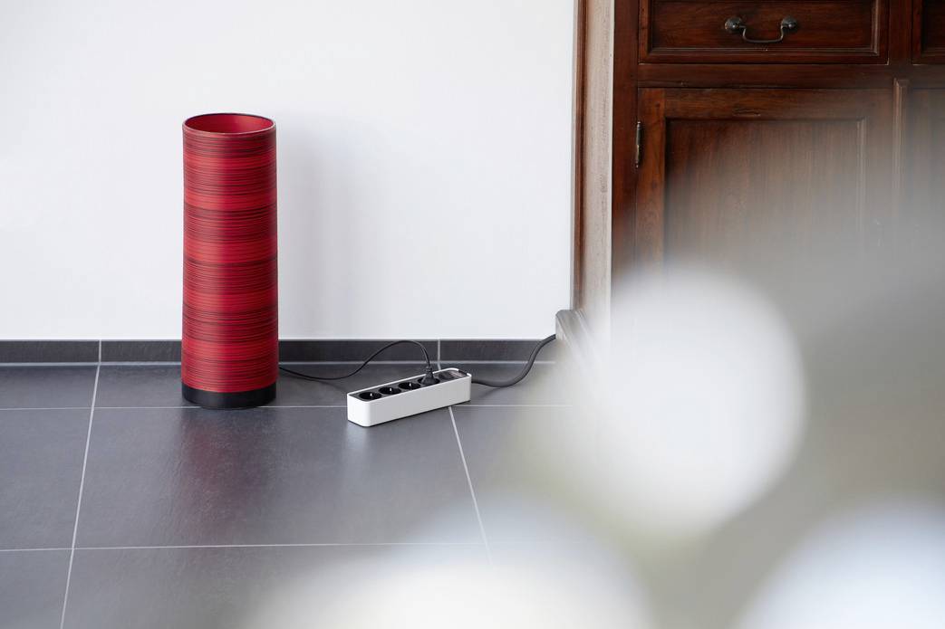 A red cylindrical air purifier stands on a tiled floor next to a power strip, in a room with wooden furniture.