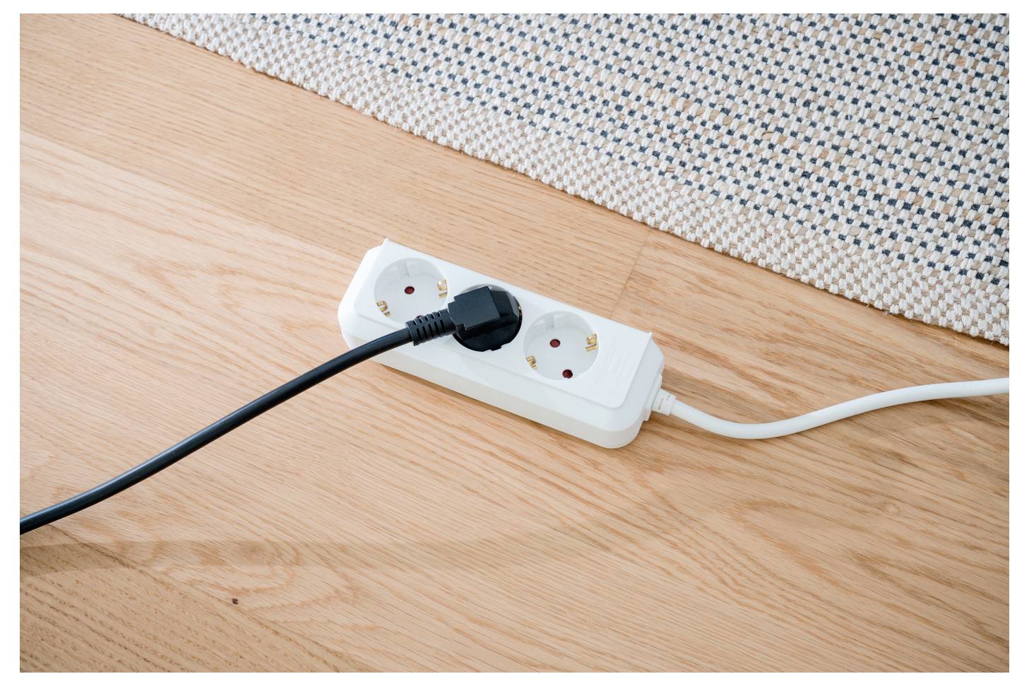 Power strip on a wooden floor with two plugged-in cords, one black and one white, against a woven rug edge.<br>