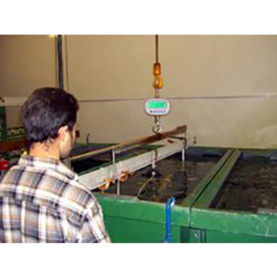 Man in a checked shirt supervising an industrial lifting procedure in a factory environment. A weight is hanging from a hook above a tank.