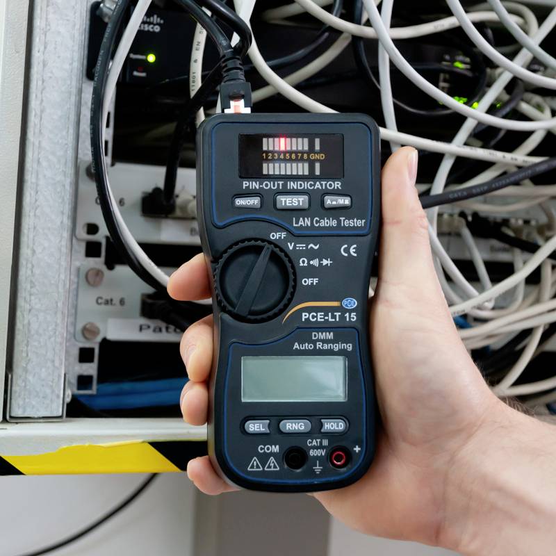 A hand is holding a LAN cable tester device connected to a network at a server rack. The screen displays test results.