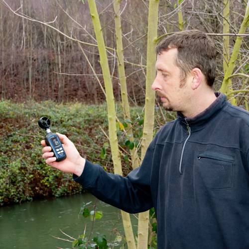 A man is measuring the wind speed outdoors near a river with a handheld device, surrounded by bare trees.