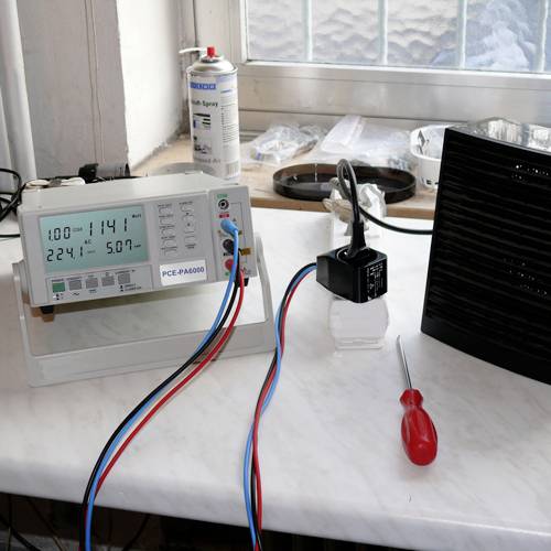 A measuring device with digital displays shows readings next to a plug socket, a screwdriver, and a spray can on a table.