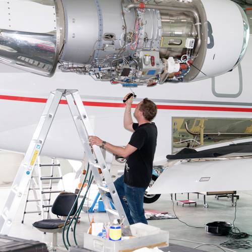 A technician stands on a ladder and inspects an aircraft engine with a torch in a hangar.