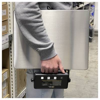 Person holding a large silver laptop against their torso in a warehouse aisle, with a smaller black device attached at the bottom.