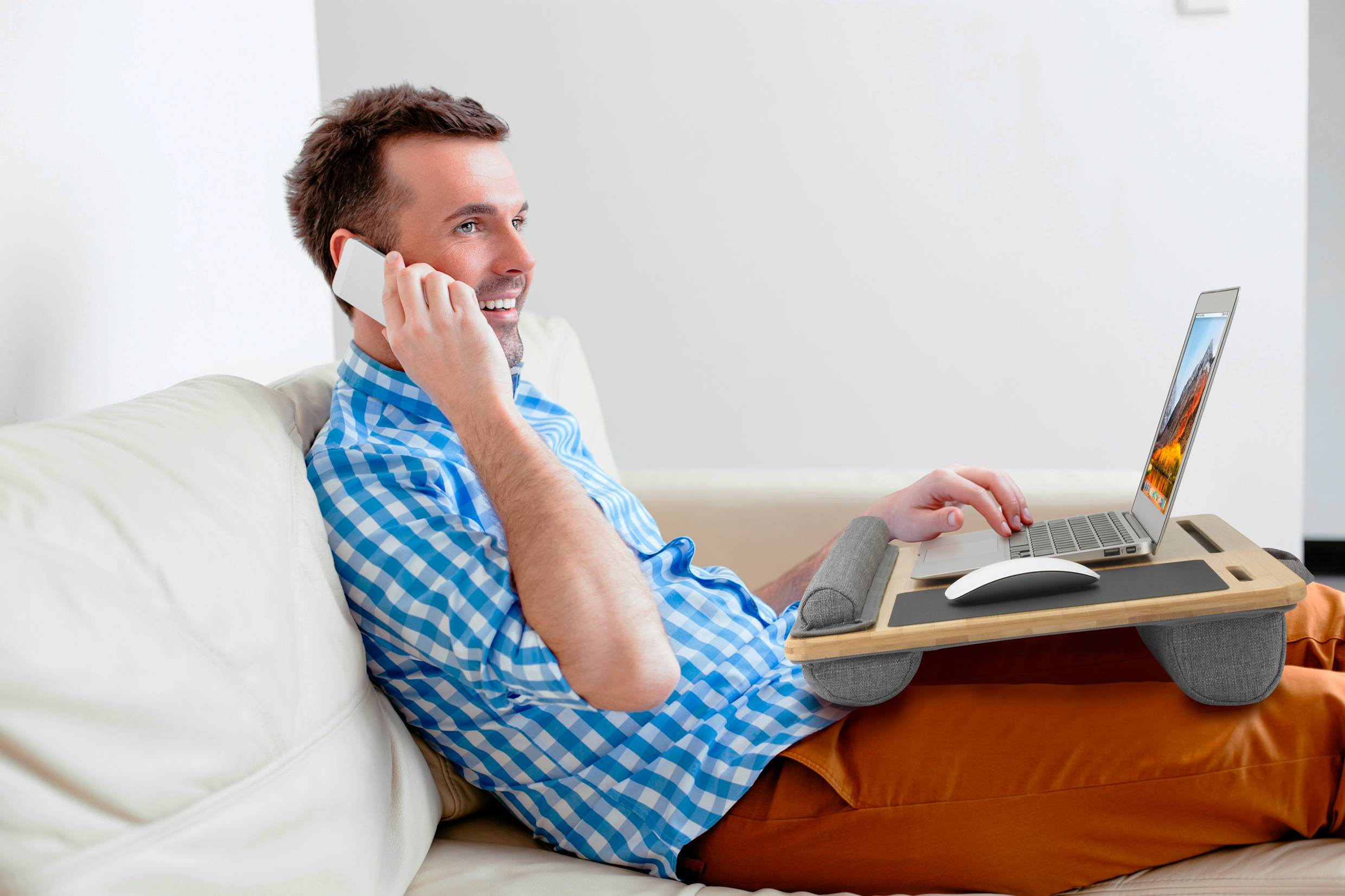 A man is sitting on a sofa and talking on a mobile phone. He has a laptop on his lap and looks content.