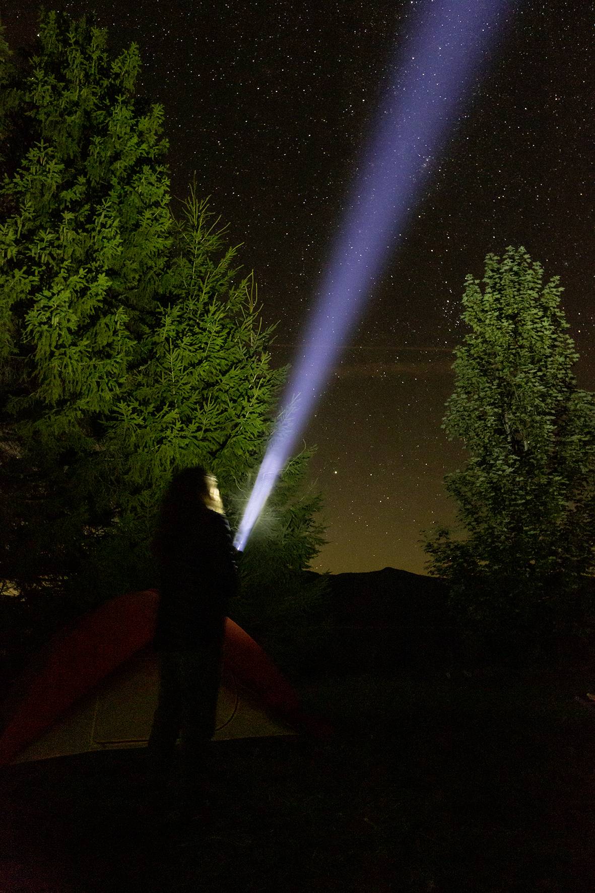 A person in front of a tent in the forest shines a torch up at the starry sky.<br><br>Note: I've made typical British English adjustments such as using "torch" instead of "flashlight" and maintained the original descriptive scene.