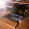 A laptop on a wooden table in an office, screen switched off. Cable plugged in. Bookshelf with books in the background.