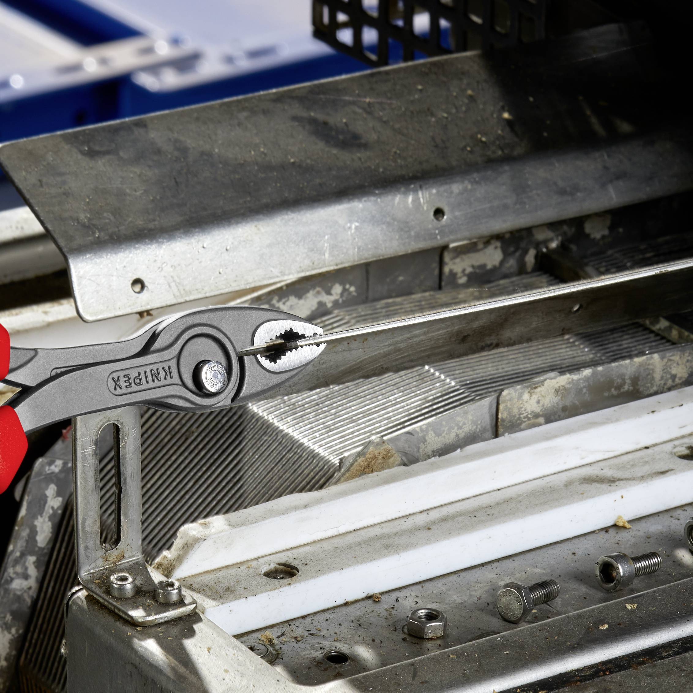 A red pipe wrench pulls on a metal sheet of a machine, surrounded by screws and technical details in an industrial environment.
