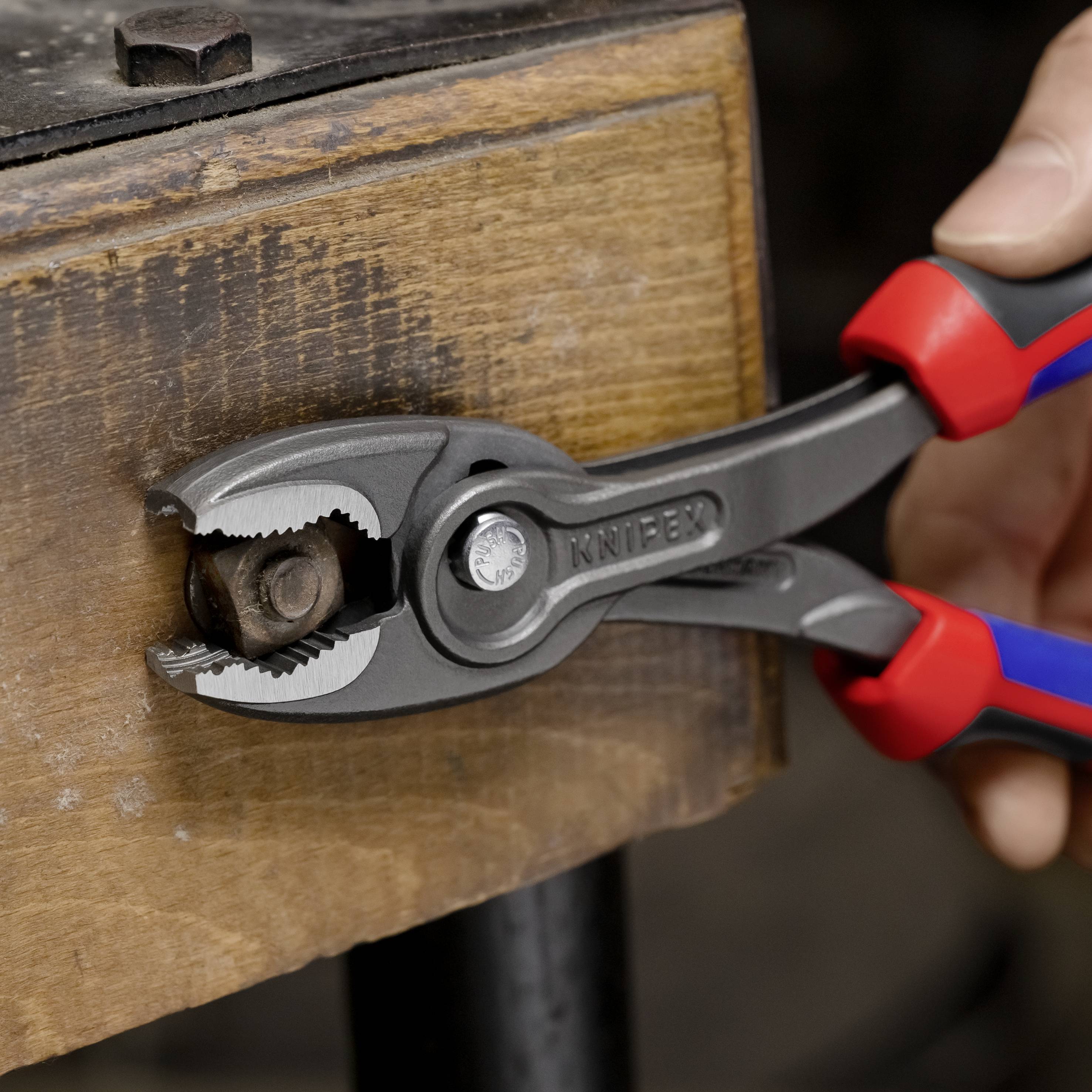 Pliers with red handles grip a screw on a wooden beam. A hand is holding the pliers.