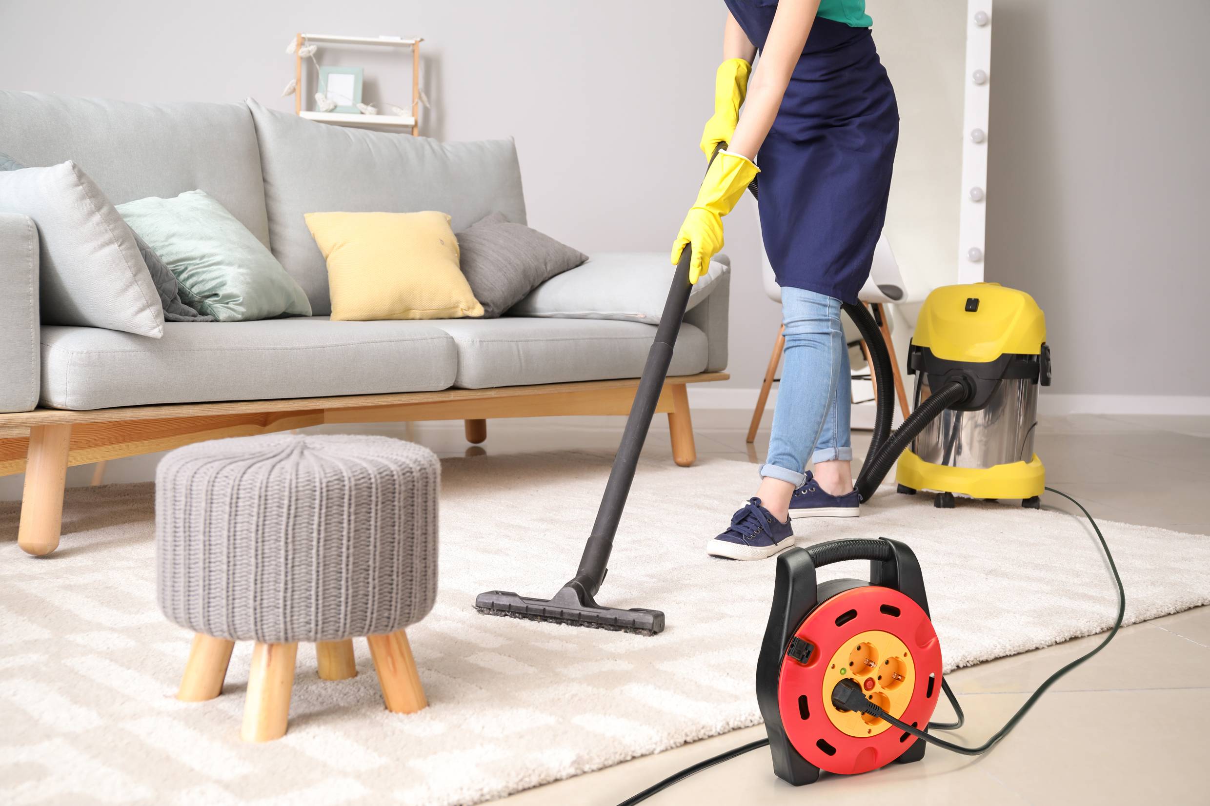A person is vacuuming a cream-coloured carpet in a living room with a vacuum cleaner. In the background, there is a grey sofa and yellow cushions.
