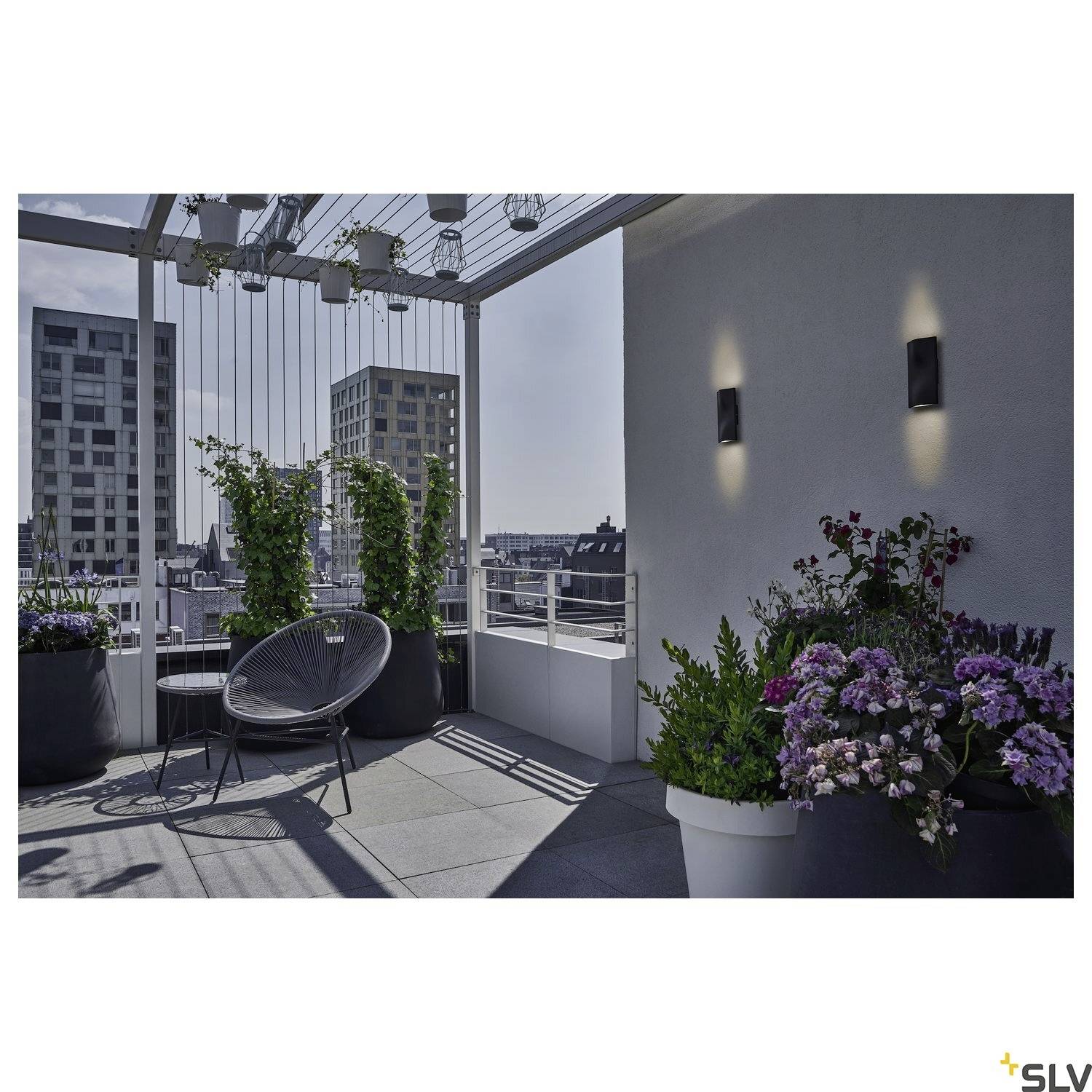 A contemporary roof terrace with plants in large pots, a black chair and wall lighting. View of urban high-rise buildings in the background.