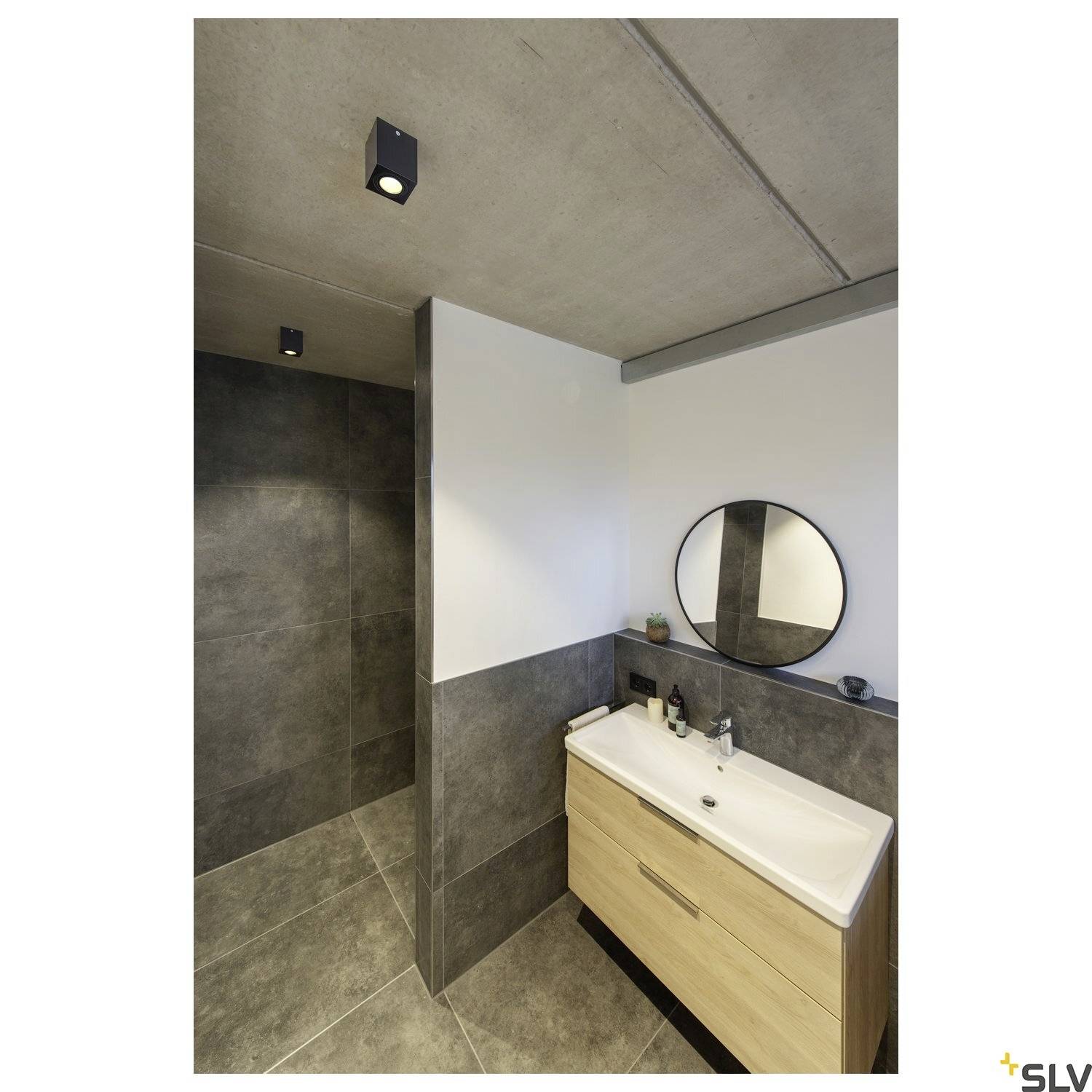 Modern bathroom with a dark, tiled shower wall, a wooden washstand with two basins and a round mirror.