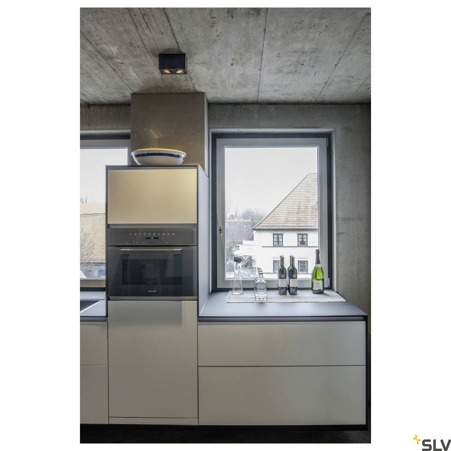 Modern kitchen with grey concrete flooring, white cabinets and built-in electrical appliances. Through the windows, a neighbouring house is visible.