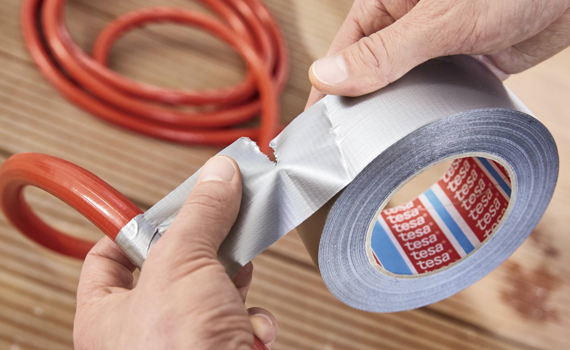 A hand tears a piece of grey adhesive tape from a reel. A red hose is visible in the background.