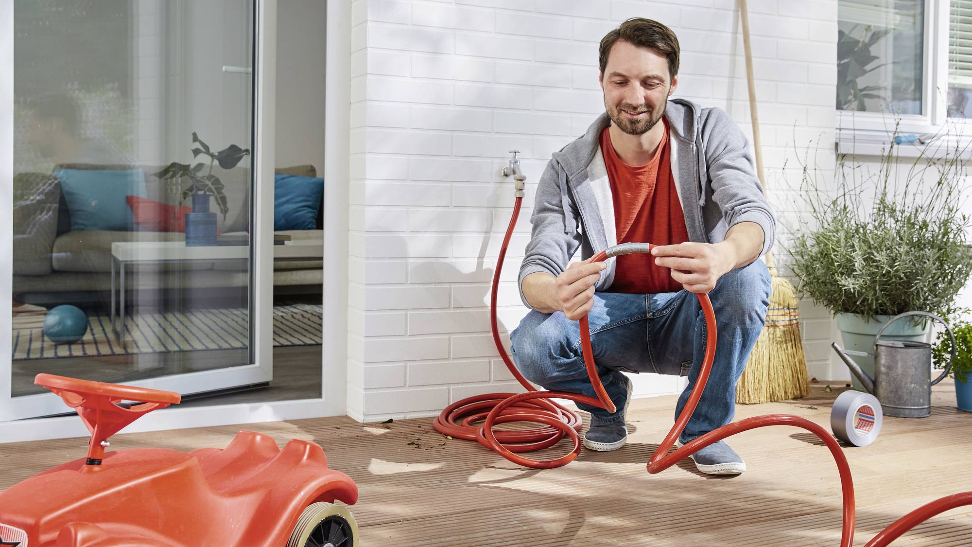 A man is kneeling on a patio and repairing a red garden hose. A front door and plants are visible in the background.