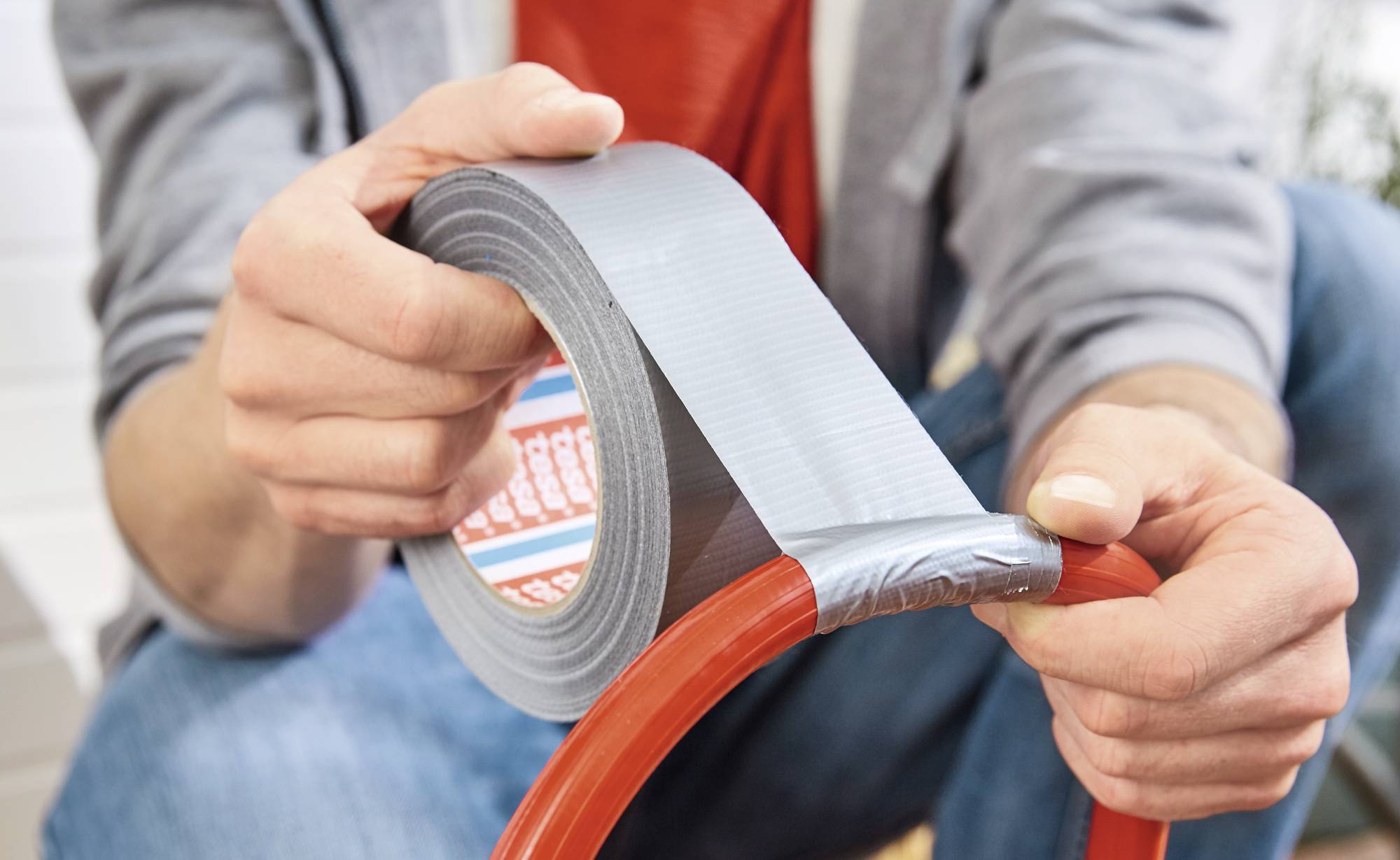 A person is wrapping silver duct tape around a red hose. Hands are holding the tape and hose firmly. The focus is on the repair.
