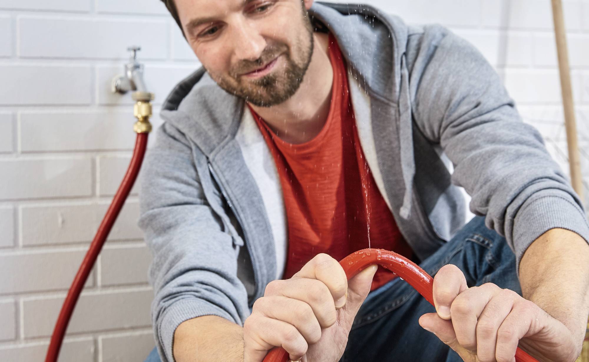 A man is repairing a red water hose in front of a white brick wall. A tap is visible in the background.