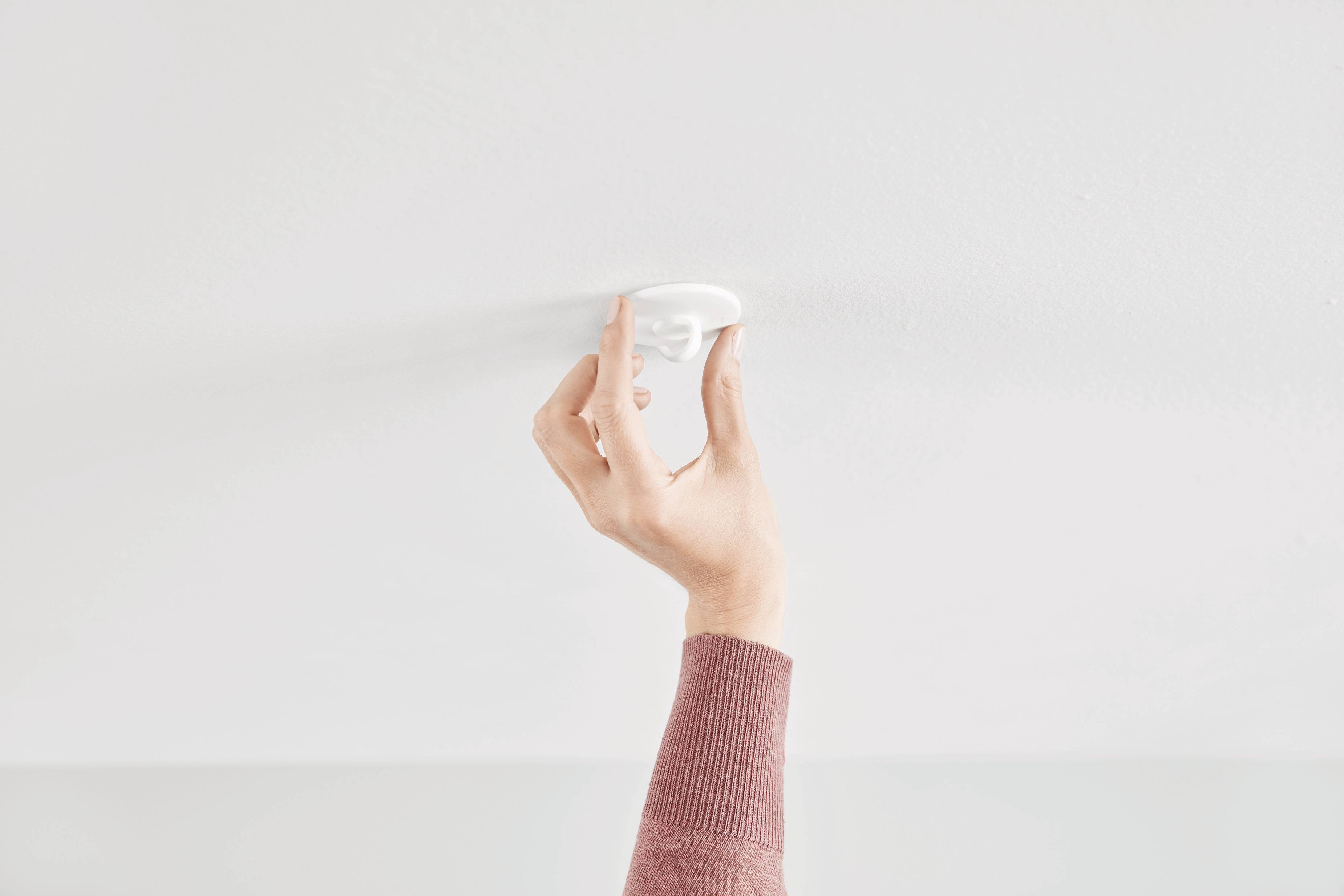 A hand in a red jumper tests a smoke detector on a white ceiling.
