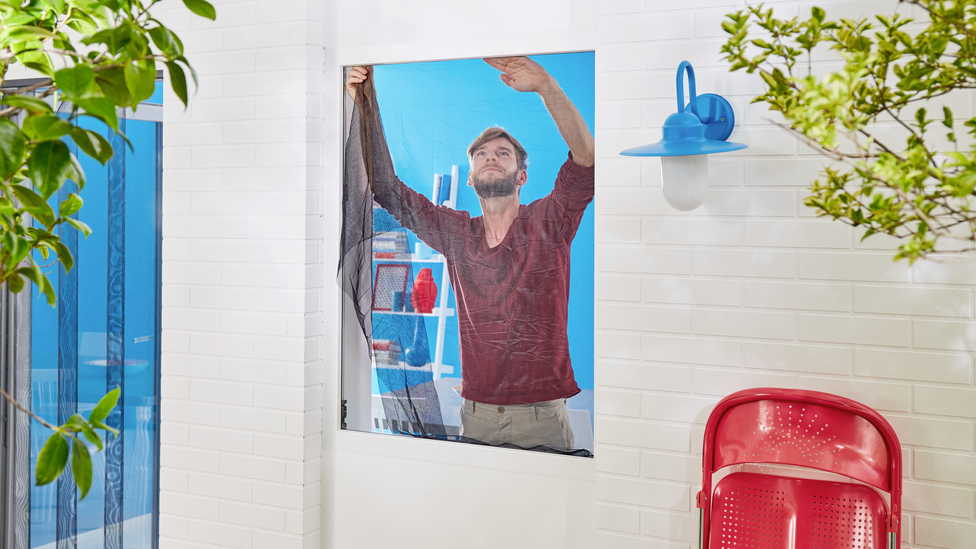 A person is securing a mosquito net to a window. Blue walls and red shelves are visible in the background.