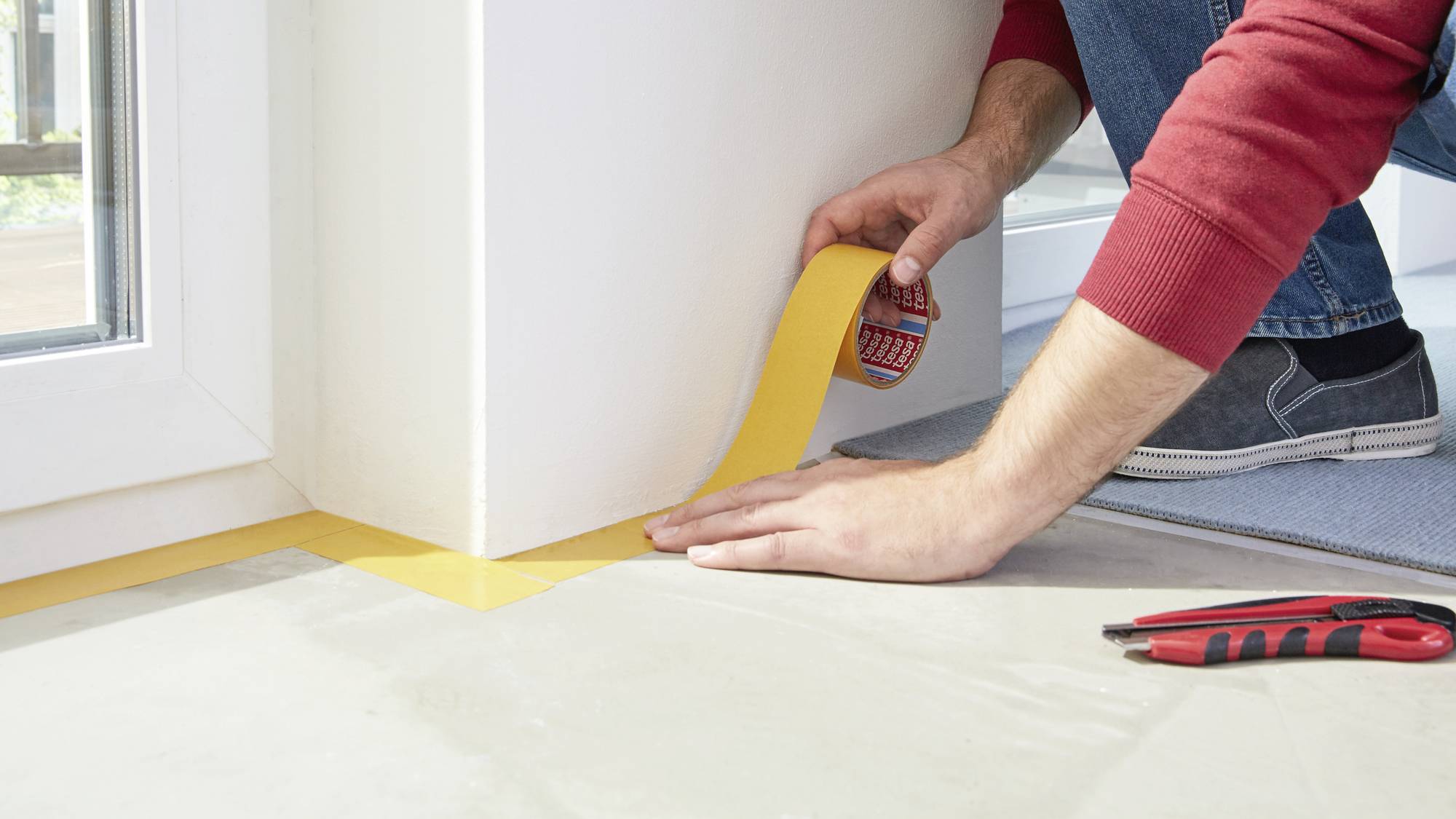 A person is applying yellow masking tape along an edge on a floor to protect walls when painting. A red and black tool lies nearby.