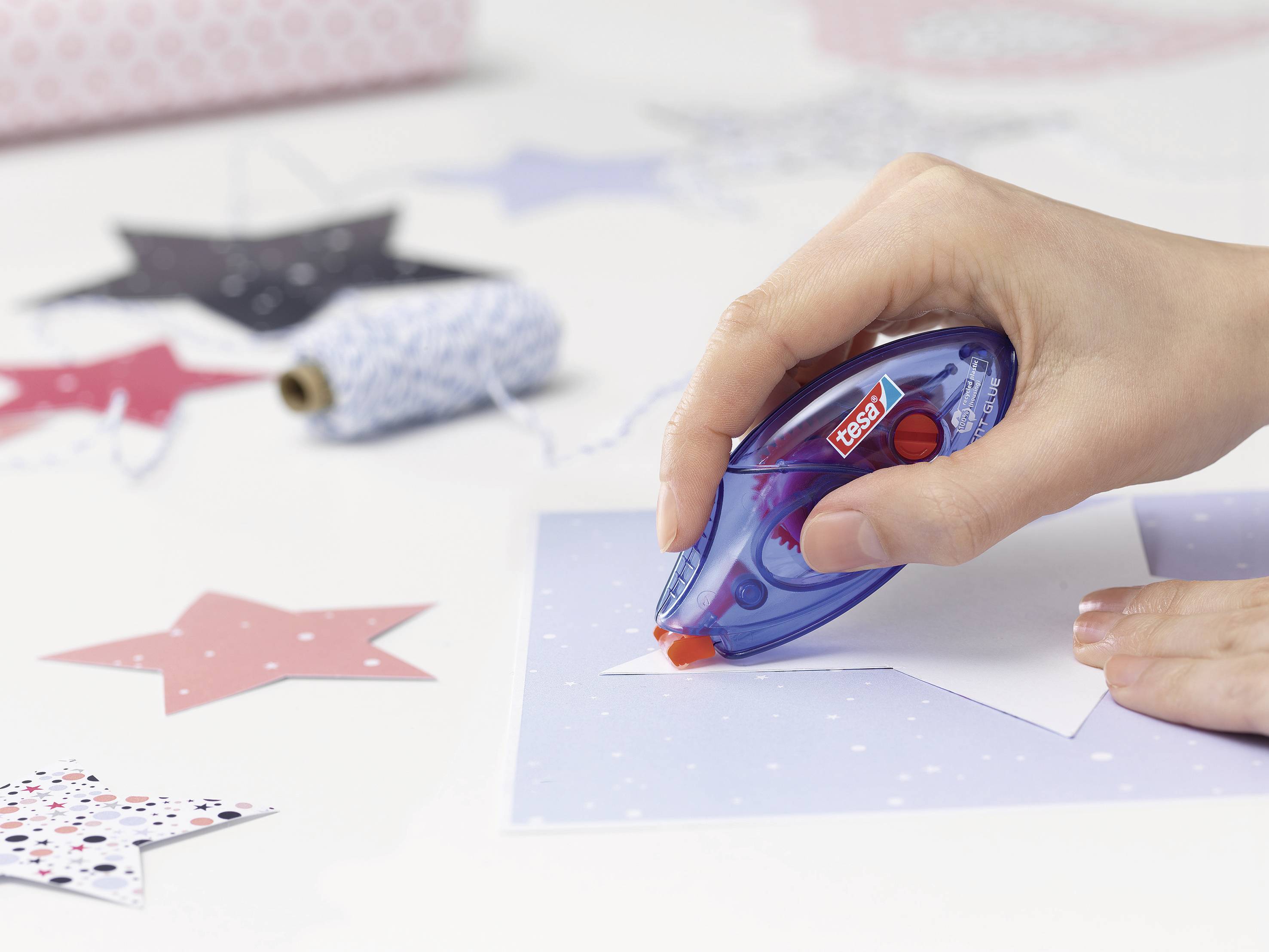 A hand is using a blue adhesive roller to stick white paper stars onto a blue sheet. Background with decorative elements.