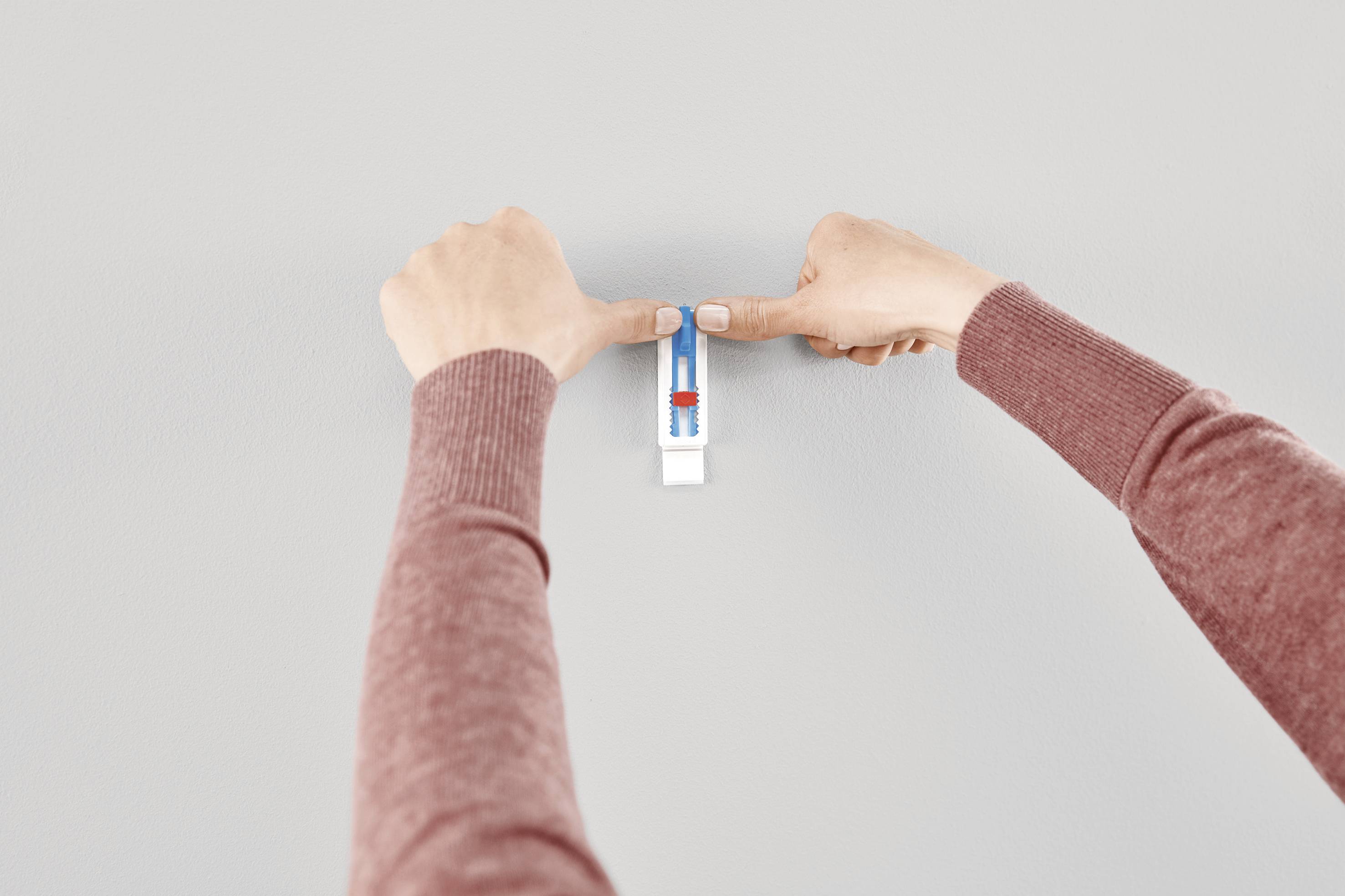 Two hands are hanging a white clip with a small spirit level on a grey wall to ensure precise alignment.