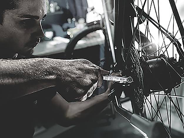 A person is repairing a bicycle, focusing on the rear wheel with a wrench in a dimly lit workshop.