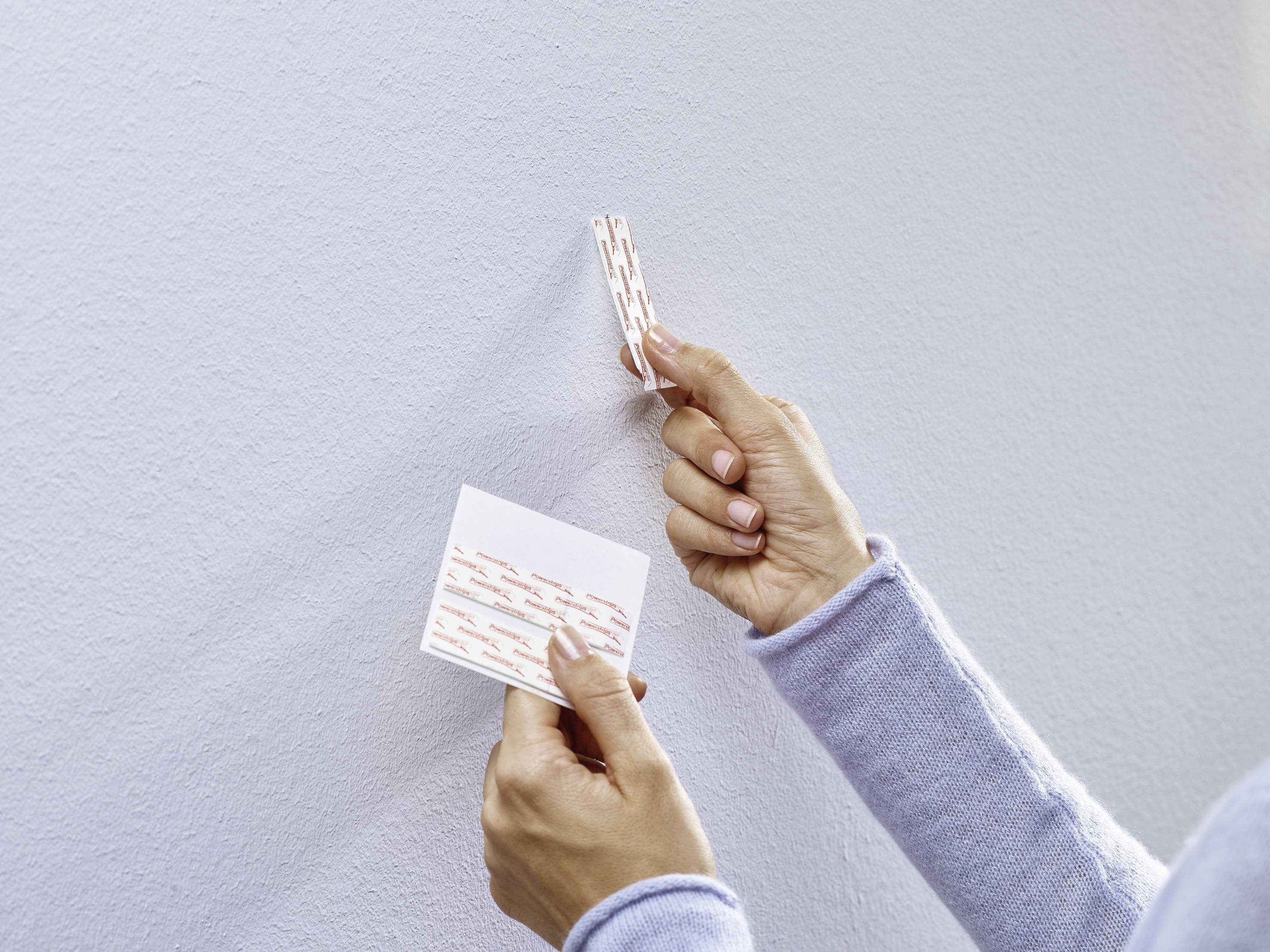 Two hands are securing a hook to a light grey wall using a double-sided adhesive pad.