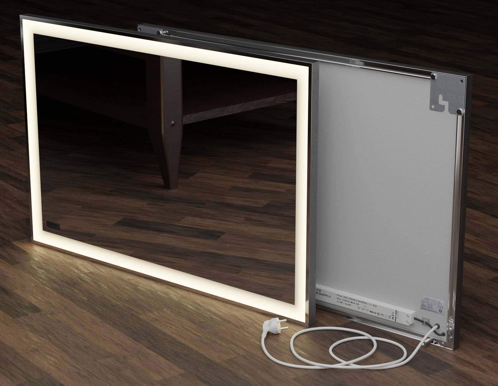 A illuminated mirror with a rectangular frame and mains plug is lying on a wooden floor and shows a table in the background.