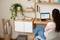 A woman is sitting at a desk with a laptop. She is working in a cosy room with plants and shelves in the background.